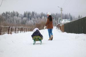 ung mor rullar henne liten söt döttrar på en kälke i vinter- dag foto
