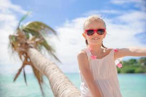 förtjusande liten flicka Sammanträde på handflatan träd under sommar semester på vit strand foto