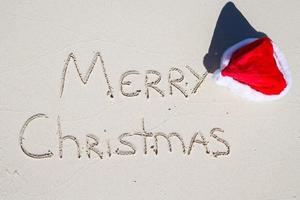 glad jul skriven på tropisk strand vit sand med xmas hatt foto