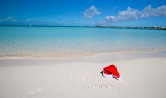 glad jul skriven på tropisk strand vit sand med xmas hatt foto