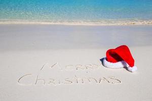 glad jul skriven på tropisk strand vit sand med xmas hatt foto