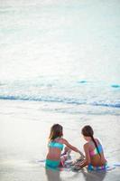 förtjusande liten flickor spelar med sand på de strand. tillbaka se av barn Sammanträde i grund vatten och framställning en korthus foto