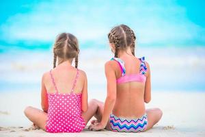 förtjusande liten flickor spelar med sand på de strand. tillbaka se av barn Sammanträde i grund vatten och framställning en korthus foto