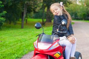 förtjusande söt liten flicka i läder jacka Sammanträde på henne leksak motorcykel foto