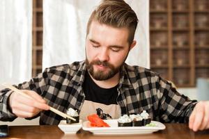 man äter sushi rullar i en restaurang. kille äter japansk kök använder sig av bambu pinnar. kultur och mat och tradition i modern värld foto