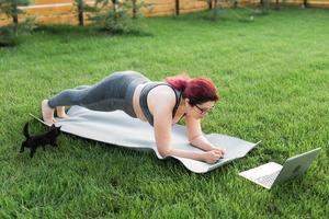 ung plus storlek kvinna i sportig topp och damasker stående i planka på yoga matta utgifterna tid på grön gräs i gård. svart kattunge promenader runt om henne. väl varelse och kondition begrepp foto