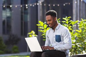 svart man frilansare arbetssätt uppkopplad använder sig av bärbar dator Sammanträde på bänk utanför kontor modern byggnad foto