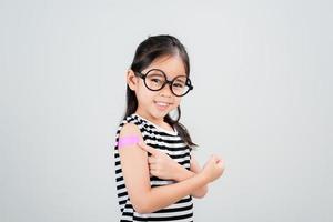 asiatisk liten flicka ha på sig glasögon som visar henne ärm med bandage efter virus vaccin Lycklig unge och barn vaccination kampanj. skydda din unge med vaccin begrepp foto