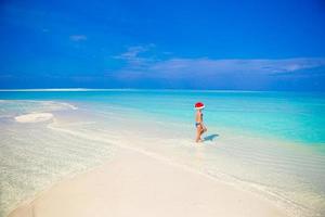 liten flicka i santa hatt på de strand under semester foto