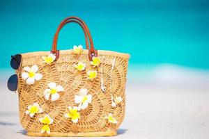 närbild vacker väska med frangipani blommor och solglasögon på vit strand foto