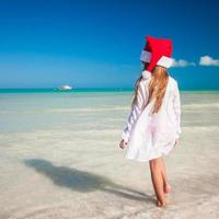 liten förtjusande flicka i röd santa hatt på tropisk strand foto