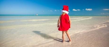 liten förtjusande flicka i röd santa hatt på tropisk strand foto