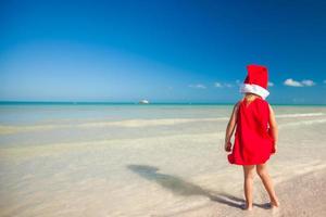 liten förtjusande flicka i röd santa hatt på tropisk strand foto
