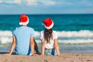 ung romantisk par i röd santa hattar Sammanträde på tropisk vit sand strand fira jul foto