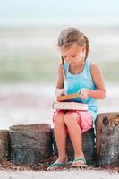 liten bedårande flicka som läser bok under den tropiska vita stranden foto