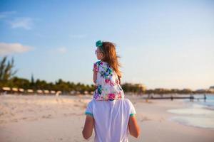 liten flicka ridning på henne pappa gående förbi de strand foto