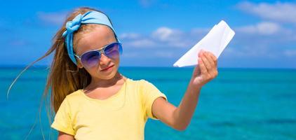 liten flicka med papper flygplan ha roligt på vit sandig strand foto