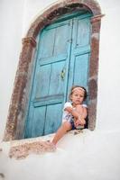 förtjusande liten flicka Sammanträde nära blå dörr av gammal hus i emporio by, santorini, grekland foto