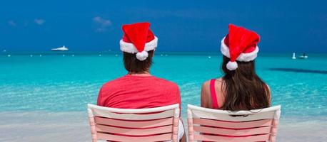 tillbaka se av ung par i röd santa hattar Sammanträde på strand stolar foto