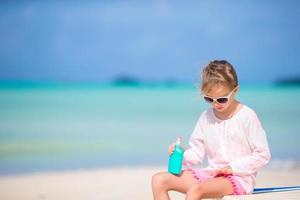 liten flicka med flaska av Sol grädde Sammanträde på tropisk strand foto