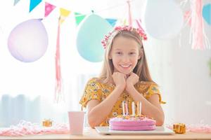 caucasian flicka är drömmande leende och ser på födelsedag regnbåge kaka. festlig färgrik bakgrund med ballonger. födelsedag fest och lyckönskningar begrepp. foto