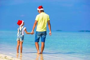 liten flicka och Lycklig pappa i santa hattar njut av strand semester foto