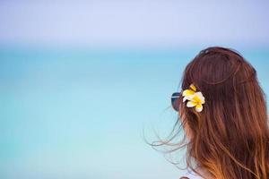 ung skön kvinna under tropisk strand semester. njut av suumer semester ensam på de strand på afrika med frangipani blommor i henne hår foto