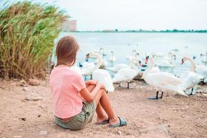 liten flicka Sammanträde på de strand med svanar foto