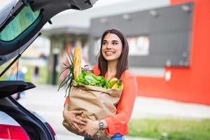 caucasian brunett gående innehav papper påsar med mat Produkter. ung kvinna sätta paket med specerier och grönsaker in i bil trunk. attraktiv caucasian kvinna handla i köpcenter eller matvaror Lagra foto