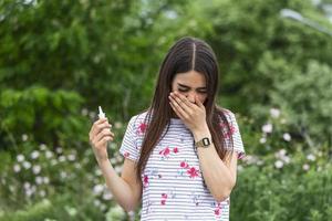 ung Söt kvinna nysning i främre av blomning träd. vår allergi begrepp. kvinna gäller nasal spray. pollen allergi symptom foto