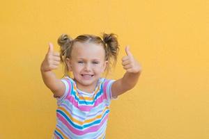 porträtt Lycklig överraskning liten unge flicka i färgad t-shirt som visar punkt fingrar tummen upp händer leende rekommendera isolerat över gul Färg bakgrund foto