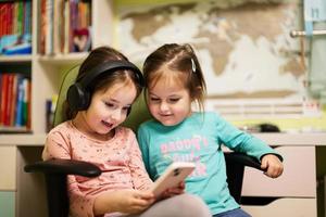 två liten flickor systrar ha på sig hörlurar tittar på tecknade serier eller unge video på mobil telefon. foto