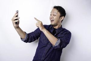 upphetsad asiatisk man bär blå skjorta leende medan innehav hans telefon, isolerat förbi vit bakgrund foto
