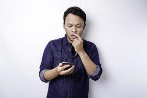 en omtänksam ung man klädd i blå skjorta medan ser åt sidan innehav hans telefon, isolerat förbi vit bakgrund foto