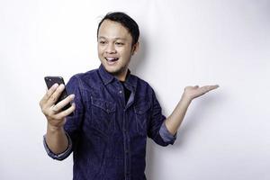 upphetsad asiatisk man bär blå skjorta pekande på de kopia Plats bredvid honom medan innehav hans telefon, isolerat förbi vit bakgrund foto