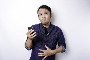 en omtänksam asiatisk man är hungrig och rörande hans mage medan innehav hans telefon tänkande Vad till ordning, isolerat förbi vit bakgrund foto