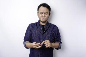 en omtänksam ung asiatisk man är bär blå skjorta innehav hans telefon och utseende förvirrad, isolerat förbi vit bakgrund foto