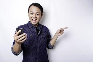 upphetsad asiatisk man bär blå skjorta pekande på de kopia Plats bredvid honom medan innehav hans telefon, isolerat förbi vit bakgrund foto