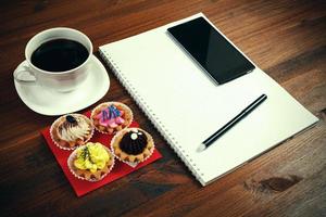 vit kaffe kopp med anteckningsbok, fyra cupcakes, smartphone och penna på en trä- tabell. foto