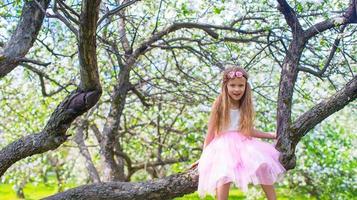 liten förtjusande flicka Sammanträde på blomstrande träd i äpple trädgård foto