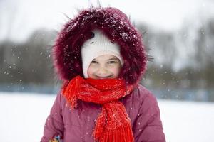 barn i vinter. en liten flicka i en värma hatt och huva utseende på de kamera och ler. foto