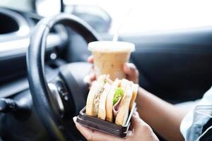 asiatisk kvinna förare håll is kaffe kopp och smörgås bröd för äta och dryck i bil, farlig och risk ett olycka. foto