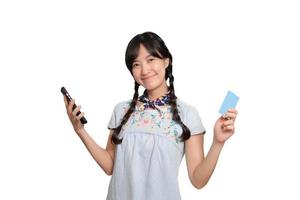 porträtt av skön Lycklig ung asiatisk kvinna i denim klänning innehav kreditera kort och smartphone på vit bakgrund. studio skott foto