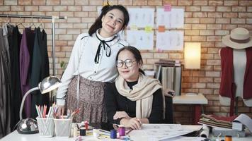 asiatisk kvinna mode designer och ung lärling se på kamera och leende i studio, vapen korsade, arbetssätt med färgrik tråd för klänning design, boutique skräddare sme företag entreprenör. foto