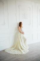 rödhårig gravid ung flicka i en vit klänning nära de vit vägg foto