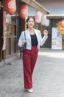 ung skön asiatisk företag kvinna ha på sig vit kostym och röd byxor leende tryggt, bär väska medan gående i de stad på en solig dag. foto