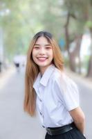 ung skön och Söt asiatisk kvinna högskola studerande bär enhetlig är stående leende lyckligt i universitet campus med träd som bakgrund. foto