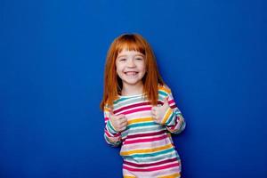 liten flicka med röd hår i en flerfärgad leende står på en blå bakgrund foto