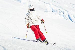 alpina åka skidor. skidåkning kvinna skidåkare gående utför mot snö täckt isolerat vit åka skidor spår backe pist i vinter. Bra rekreations kvinna skidåkare i vit åka skidor jacka och röd byxor foto