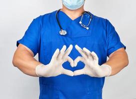 läkare i blå enhetlig och latex handskar visar med två händer en hjärta gest foto